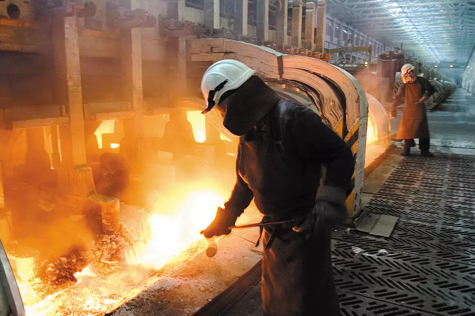 Operário com equipamento de proteção realizando a limpeza em área industrial com altas temperaturas e chama intensa ao fundo.